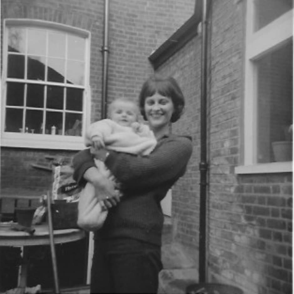 Kate’s mother as a young woman, laughing, holding Kate as a baby in the yard of a brick house
